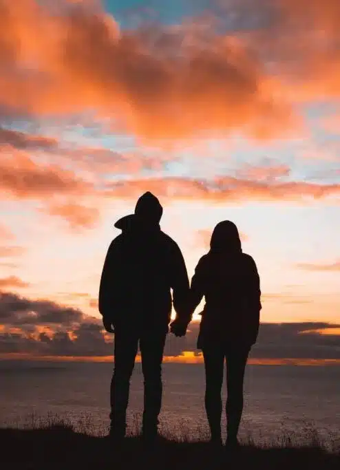 silhouette of man and woman holding hands and looking at a sunset over water